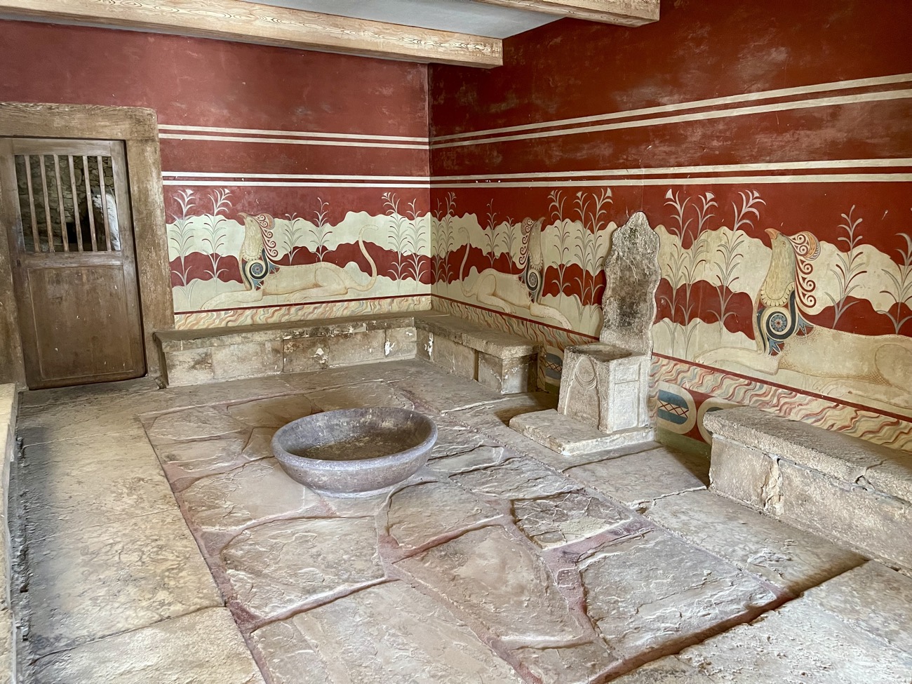 The Throne Room at Knossos — a stone throne set against the wall, flanked on both sides by painted griffins (composite lion-and-bird creatures) reclining amid stylized reeds, with gypsum benches and a stone basin on the floor