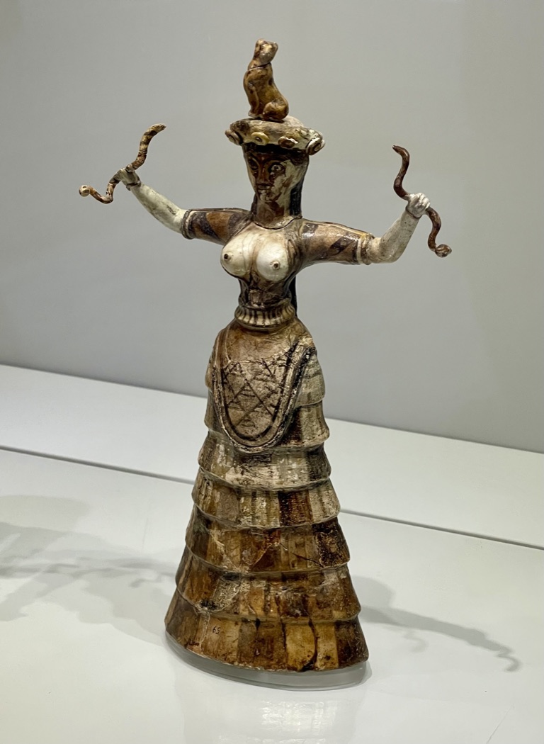 Minoan faience figurine of a goddess holding a raised serpent in each hand, with bared breasts and a tiered skirt, a feline crowning her head